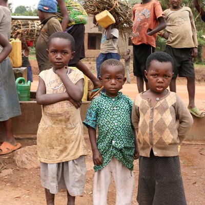 Drei Kinder mit ernsten Gesichtern im Vordergrund – im Hintergrund Menschen mit Kanistern und Brennholz beim Wasserholen.