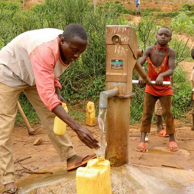Mann füllt einen Kanister, während ein Kind an der Handpumpe sauberes Wasser aus einem Brunnen fördert.