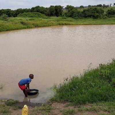 Junge Person in Ruanda schöpft braunes Wasser aus einem Gewässer mit einer Schüssel.